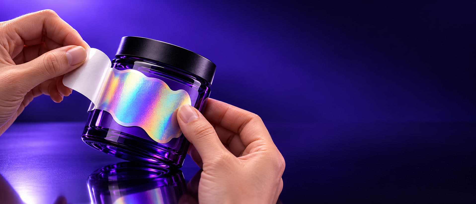 Hands peeling the backing from a holographic label and applying it to a purple glass jar on a reflective black surface