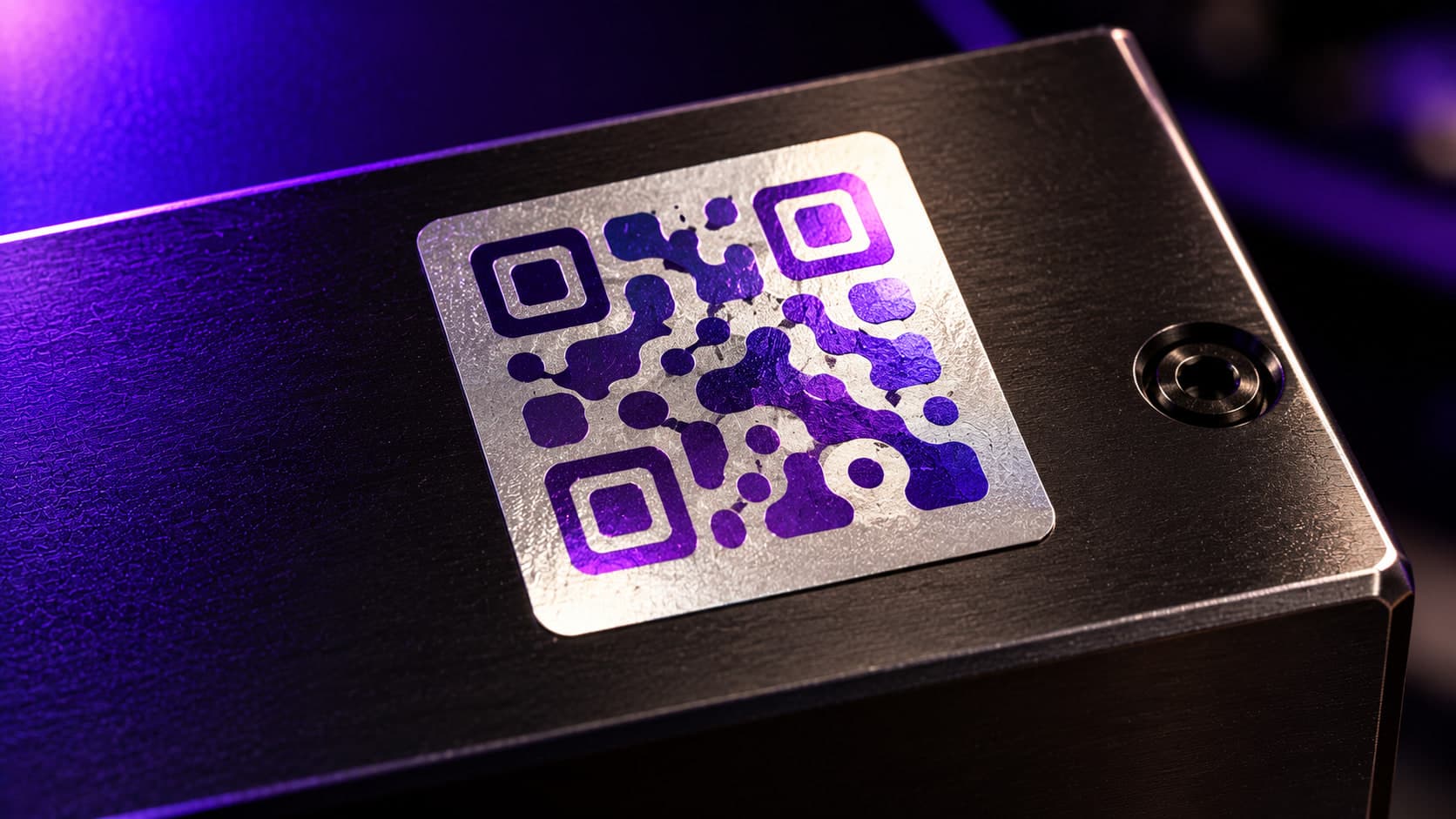 A silver polyester asset label with a violet QR block applied to a dark machined-metal housing